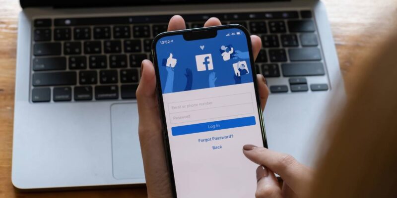 Close up of a person's hand holding a smartphone with the Facebook app login page and a laptop in the background.